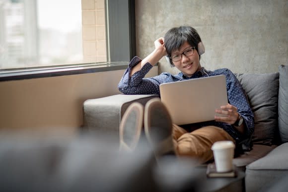 Young Asian man relaxing on a couch with his laptop and headphones.