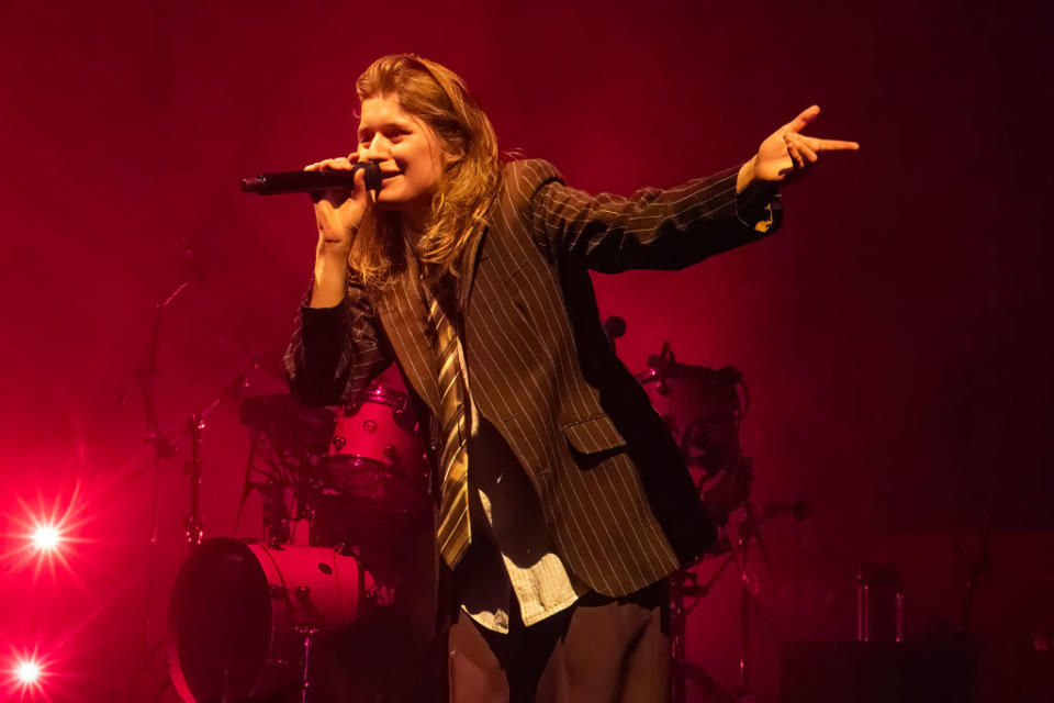 girl in red on stage performs in striped blazer and shirt, gesturing with one hand, other holds microphone