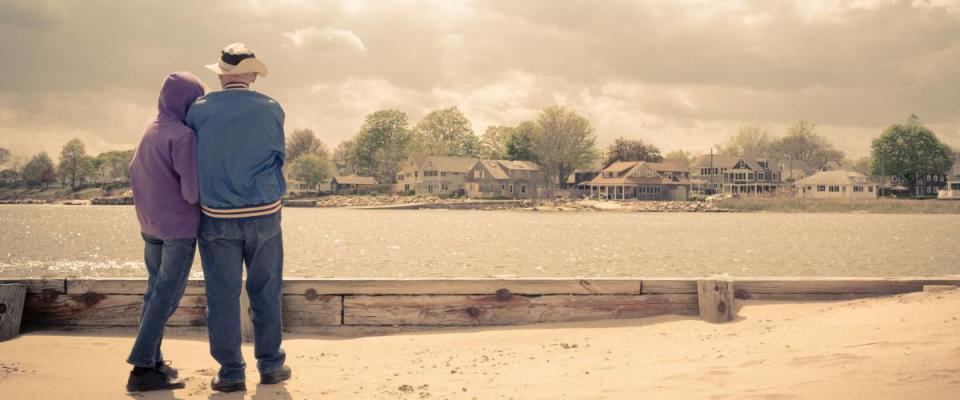 An older couple on the beach looking across the water at beautiful waterfront homes.  Vintage effect applied.