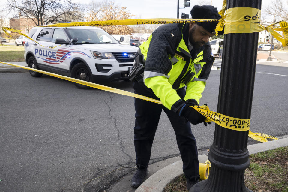 A police officer in a neon yellow jacket stands on a curb near a police vehicle and affixes yellow tape to a lightpost.