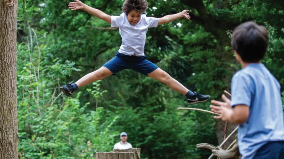 A boy doing a star jump off some play equipment in the arboretum