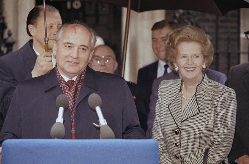 A man in a dark overcoat smiles as he speaks before microphones. With him is a smiling woman, right.