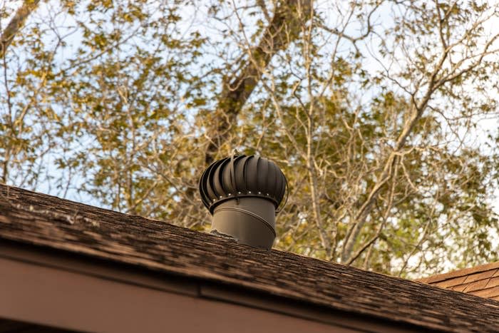 A spinning roof vent turbine is installed on a shingled roof surrounded by trees