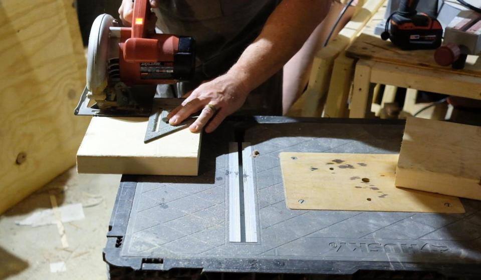 A man in a workshop using a circular saw and a speed square to make a square cut on a small piece of wood.