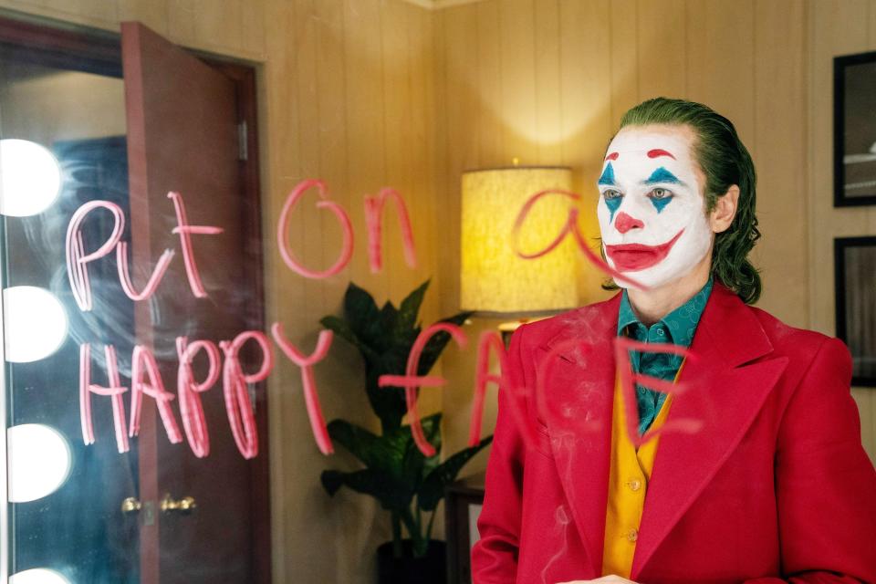 Joker from the first film standing in front of a mirror that says "put on a happy face"