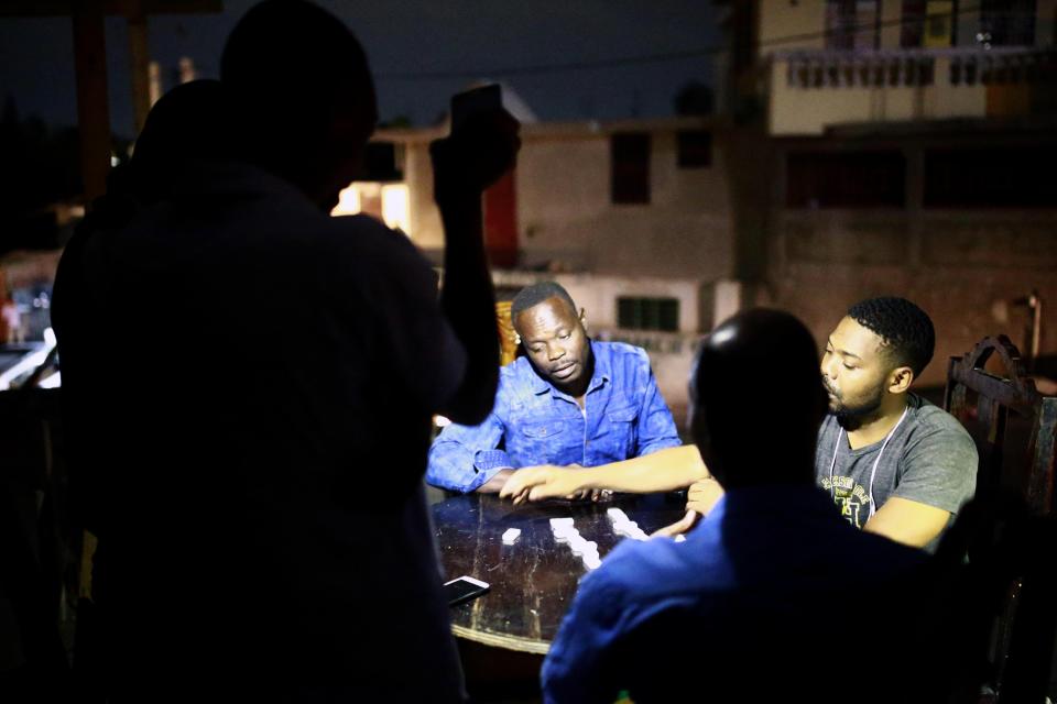 In this April 16, 2019 photo, a man holds up his mobile phone to light others playing dominos in Petion-Ville, Haiti. Nighttime activity has ground to a halt as armed robbers hold up street merchants or break into people's homes in darkness. (AP Photo/Dieu Nalio Chery)