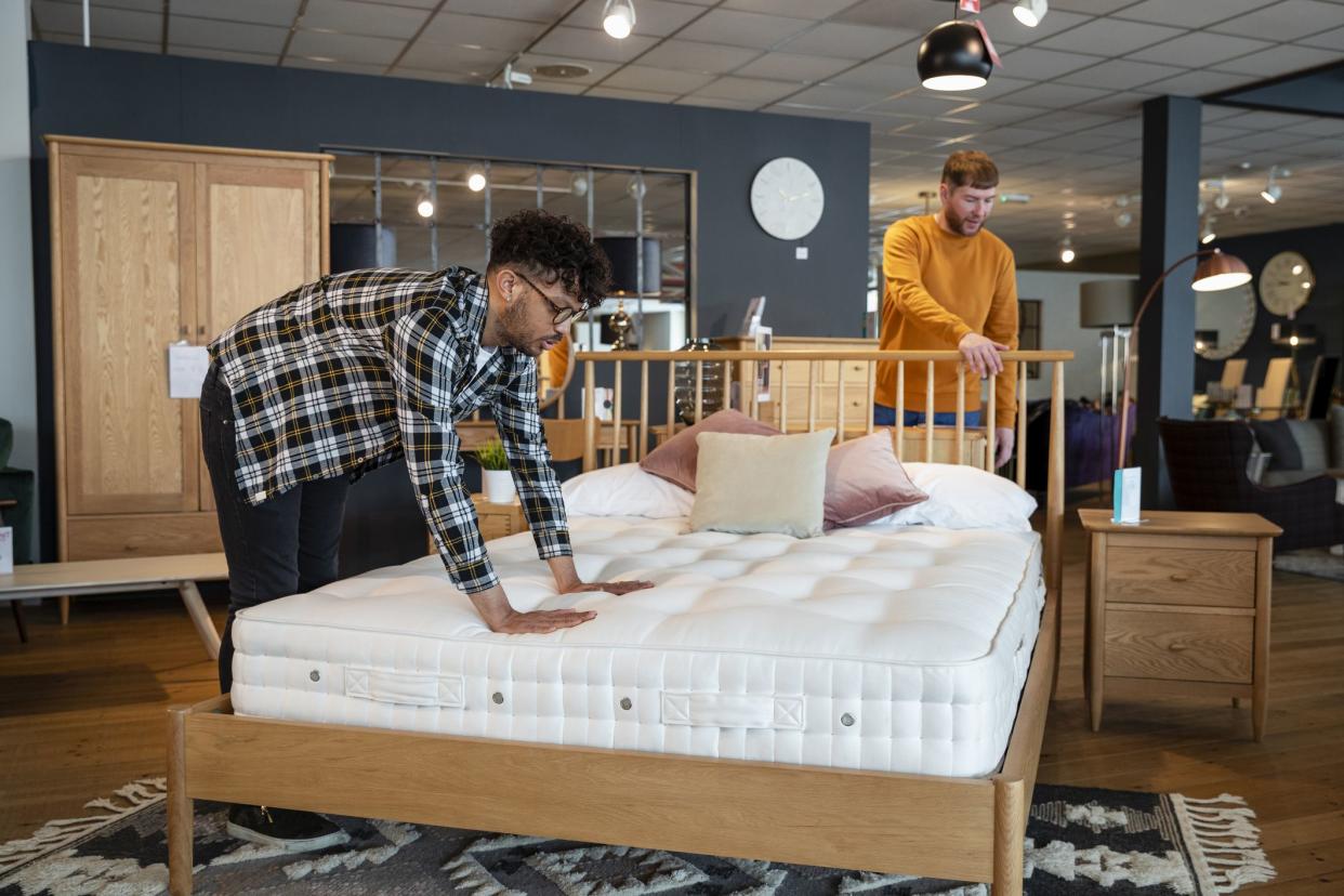 A mid adult couple viewing a home furniture showroom together, they are touching and examining a bed, considering buying it. One man is feeling the mattress while his fiancé is touching the bed frame.