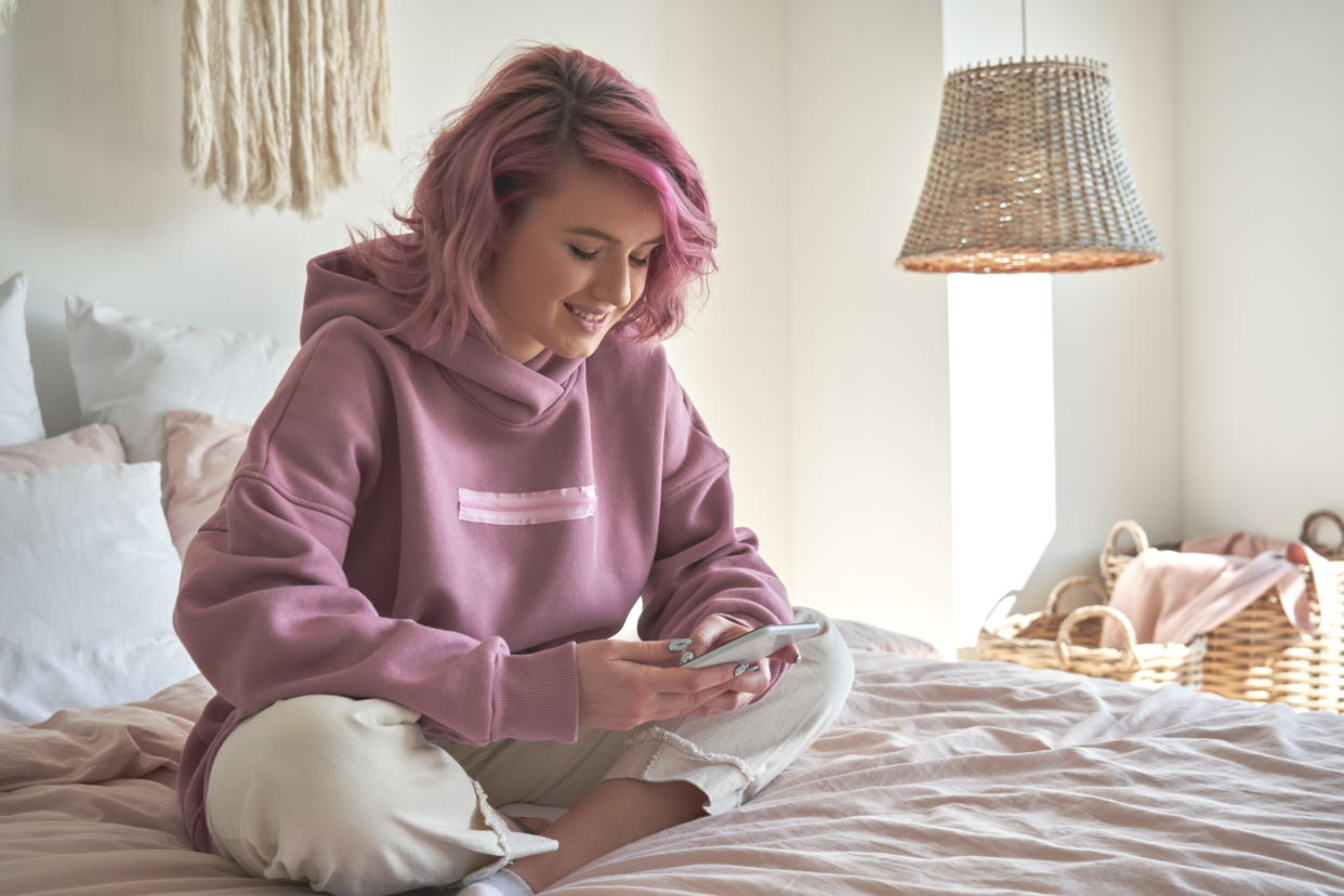 Smiling hipster teen girl with pink hair holding smart phone chatting in social media, watching video content, texting messages, playing games, using mobile apps, surfing internet sit in bed at home.