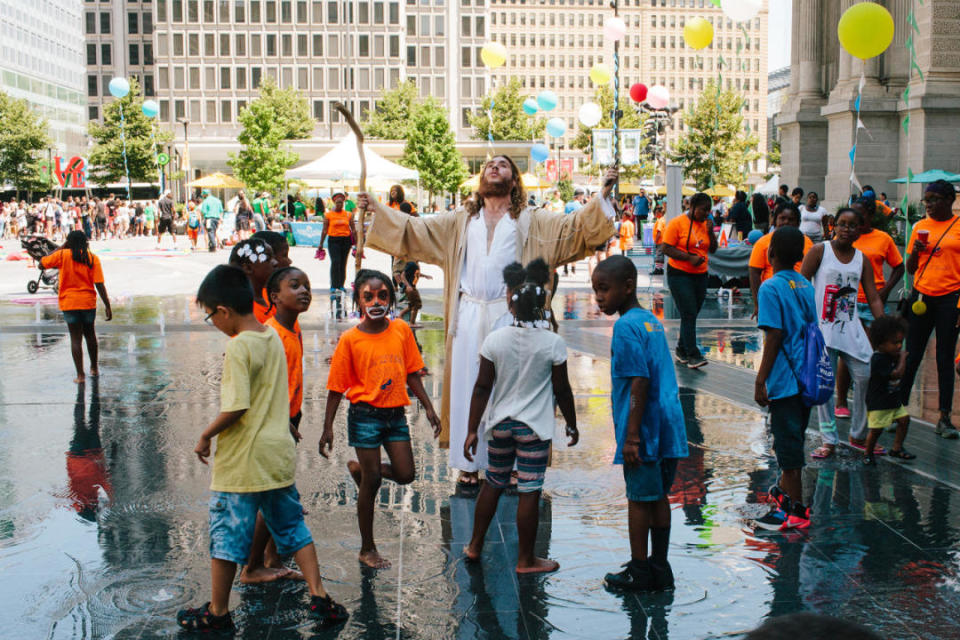 A Jesus impersonator listens to his iPhone while children play in a park in downtown Philadelphia.