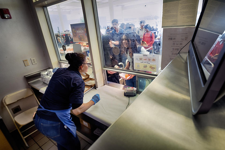 Cafeteria worker Nuria Alvarenga serves lunch to students through a service window at Firebaugh High School in Lynwood, Calif. on Wednesday, April 3, 2024. Demand for school lunches has increased after California guaranteed free meals to all students regardless of their family's income. Now, districts are preparing to compete with the fast food industry for employees after a new law took effect guaranteeing a $20 minimum wage for fast food workers. (AP Photo/Richard Vogel)