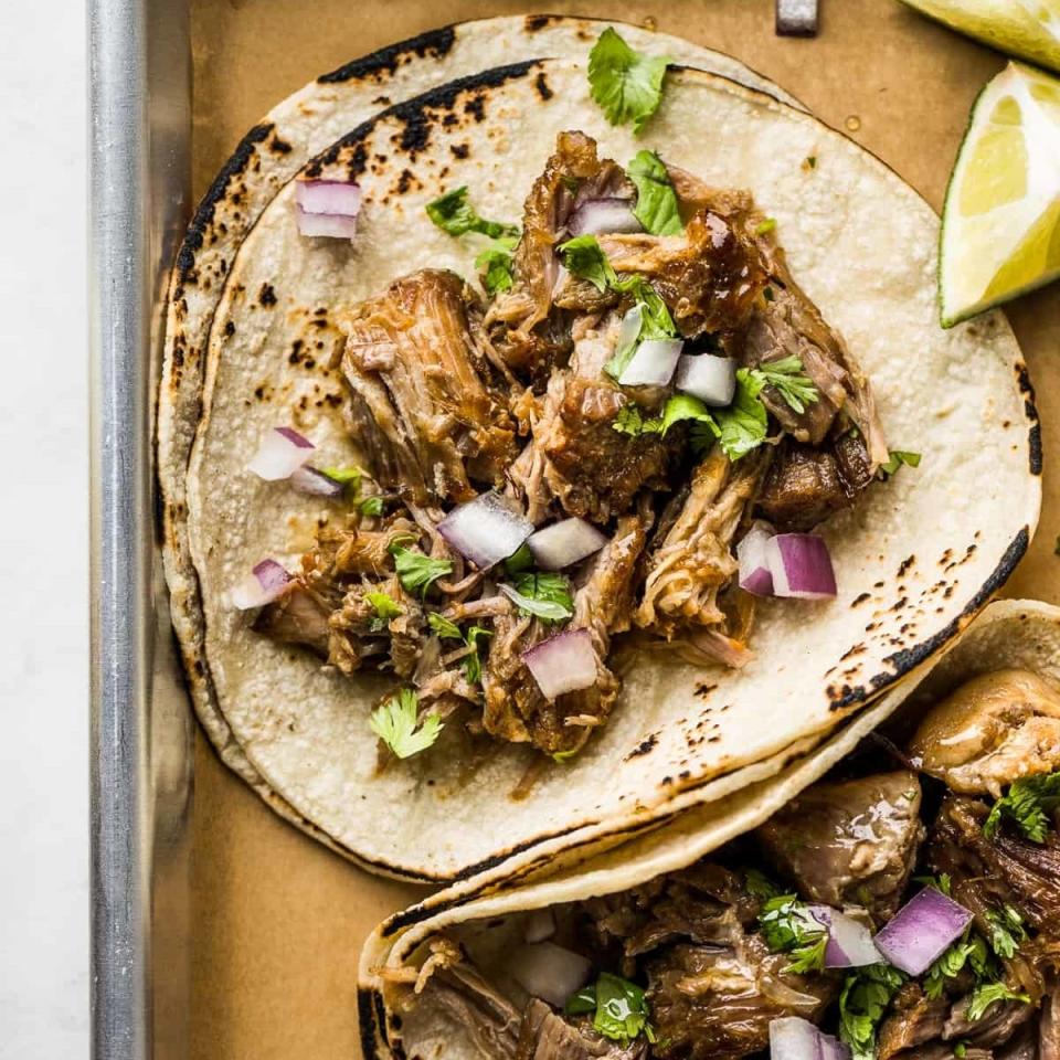 Two tacos with shredded meat and diced onions on a tray, garnished with cilantro and lime wedges