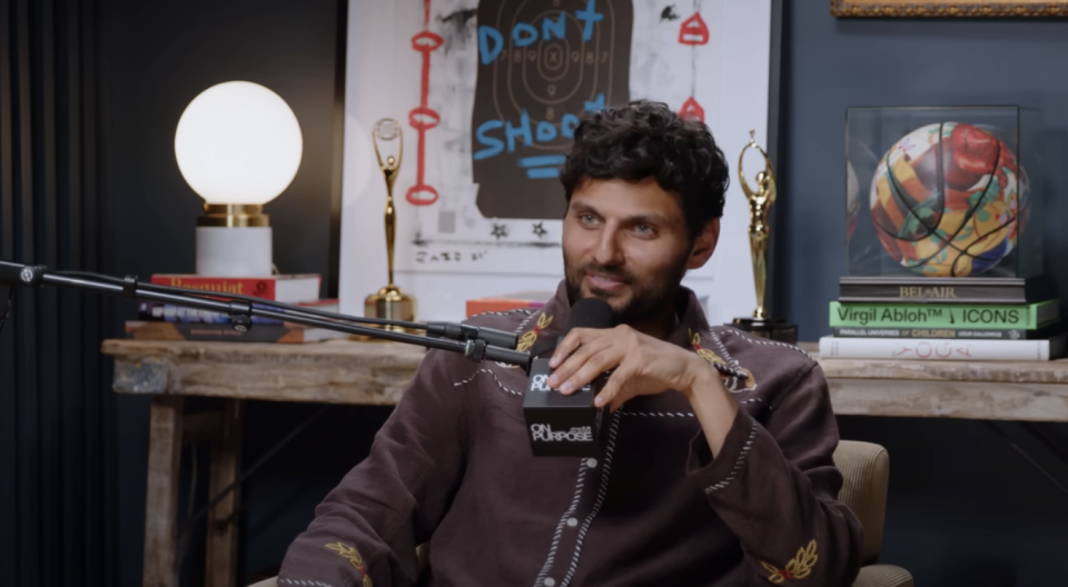 Jay sitting with microphone, wearing a zip-up top with designs, in a room with posters and a basketball