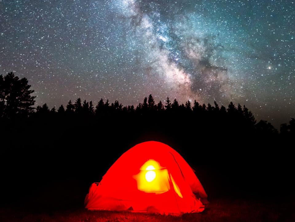 A tent is lit up from the inside so as to appear bright orange in an otherwise dark forested area. The milkyway and countless bright stars dot the sky above it.