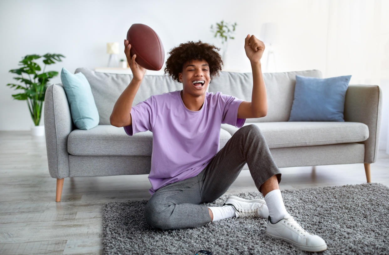 Excited teen holds football while watching TV in living room. 