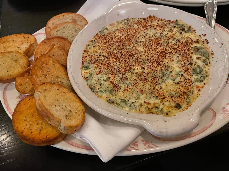 A bowl of artichoke dip with seasonings sprinkled on top sits on a plate. Small slices of bread sit on the platter next to the dip