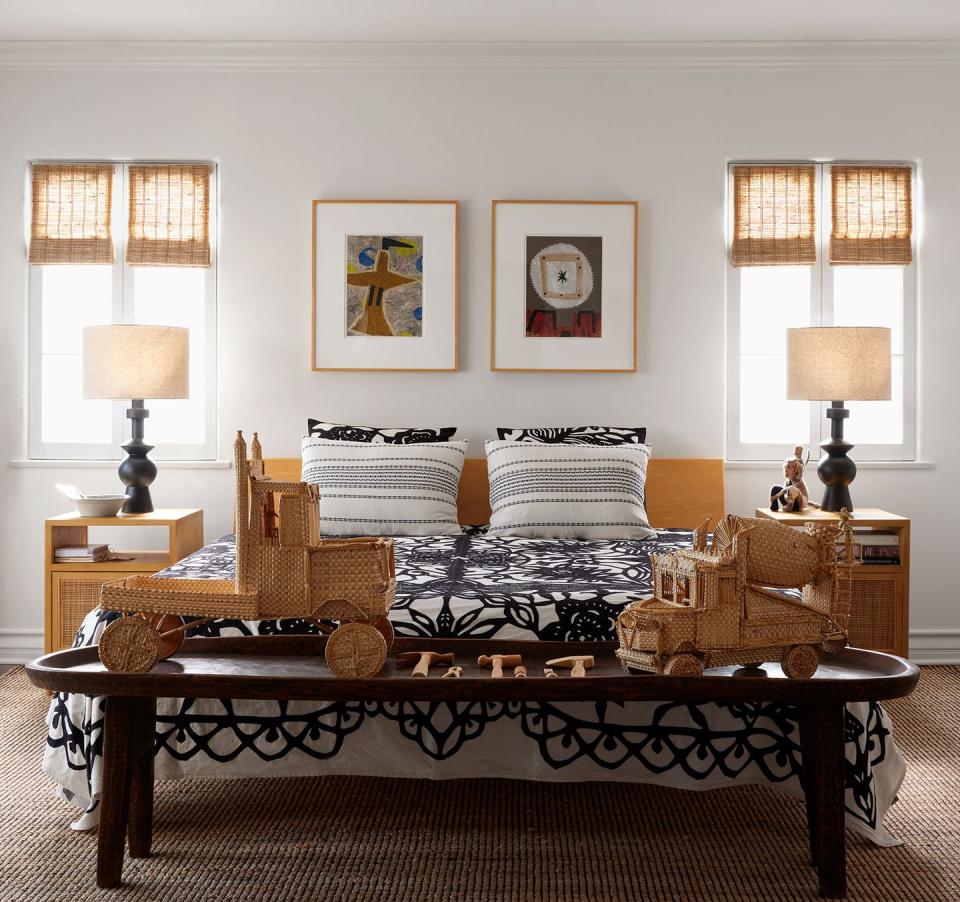 a light filled bedroom with bamboo shades has a bed with wood headboard and an embroidered moroccan coverlet, nightstands with table lamps, a wood bench at foot of bed, and wall art includes collages and a tapestry