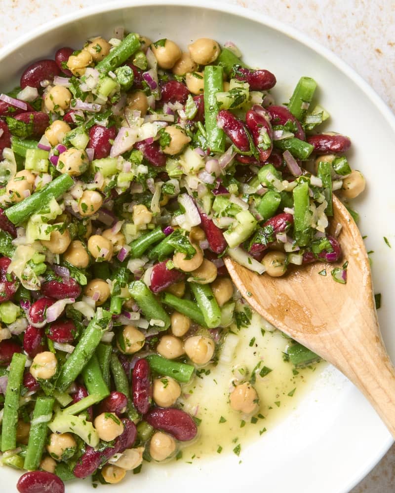 overhead shot of a 3 bean salad in a white bowl, with a wooden spoon in the bowl, with some missing from the bowl