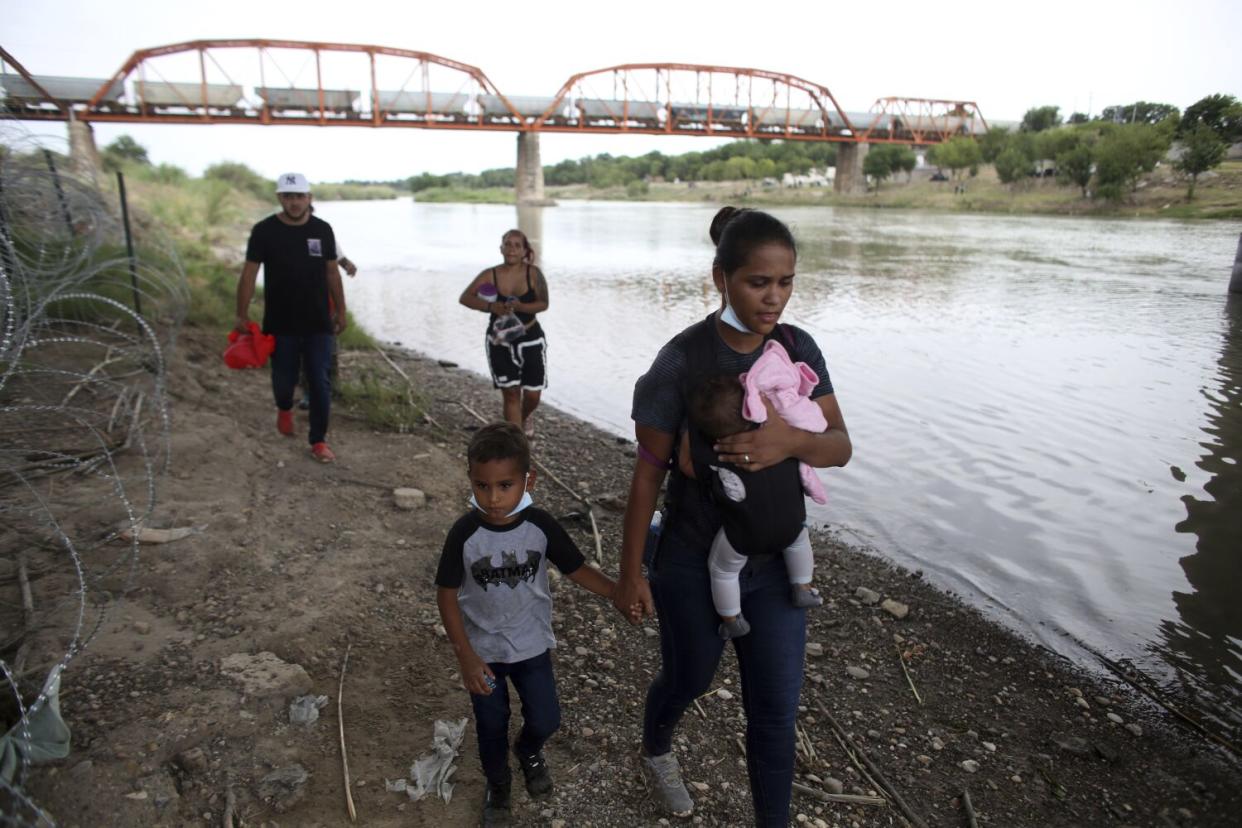 A woman carrying an infant holds the hand of a young child, while two people walk behind them along a river