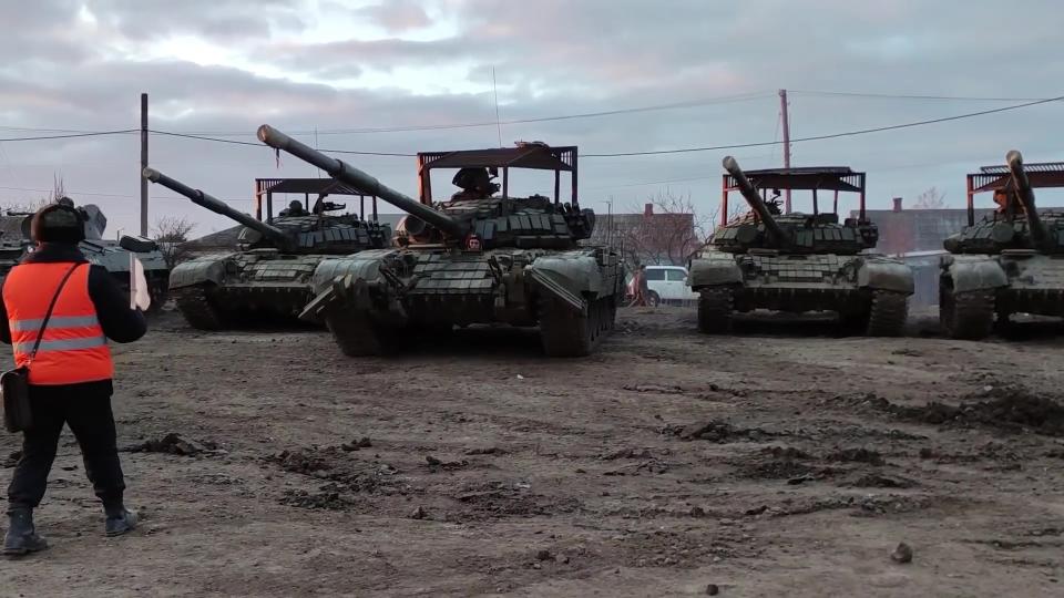 A man in an orange security jacket and helmet faces a row of Russian tanks.