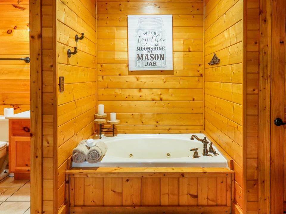 A white soaking tub inside of a wooden nook with soaps and towels on the left side, hooks on the left side of the wall, and a textual image framed in the back center wall