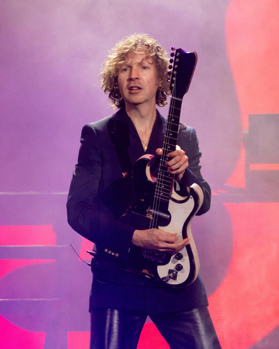 Beck performs on stage holding a guitar, wearing a dark blazer and leather pants