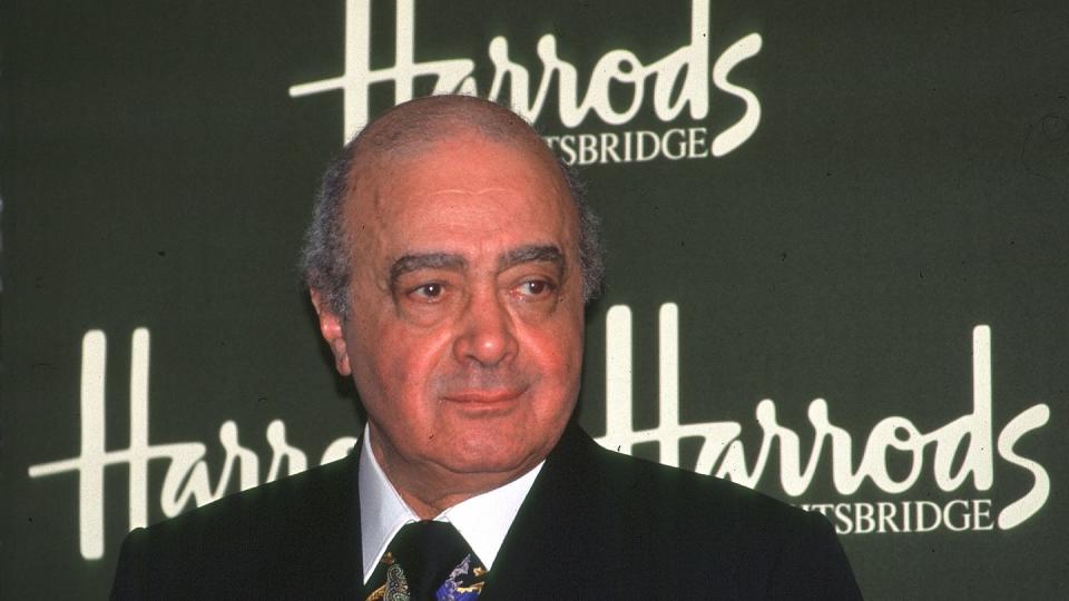 mohamed al fayed stands in front of a green harrods background, he wears a black suit jacket and tie with a multicolored collared shirt