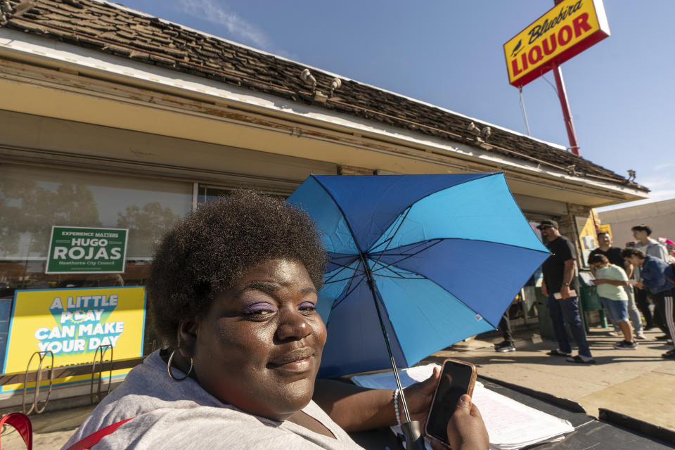 Kianah Bowman, 24, sits outside the Bluebird Liquor store collecting signatures for a petition demanding lower gas prices, as people line up to purchase lottery tickets for the Saturday drawing of the Powerball lottery at the Bluebird Liquor store in Hawthorne, Calif., Saturday, Nov. 5, 2022. Bowman has yet to buy any lottery tickets. (AP Photo/Damian Dovarganes)