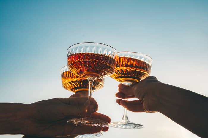 Hands holding three vintage-style glass cups filled with a sparkling beverage raised in a toast against a clear sky