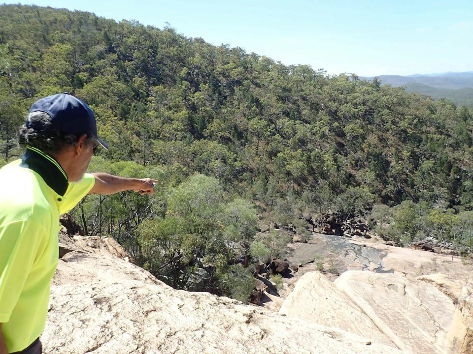 Rear view of a man wearing a hi-vis shirt pointing over a rock ledge into a valley.