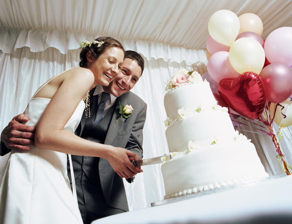 A bride in a white sleeveless dress and a groom in a gray suit, smiling and cutting a three-tier wedding cake together