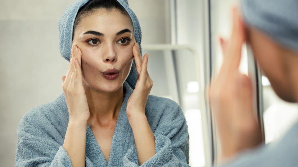 Woman in a bathrobe and towel, likely after a shower, looking in the mirror while applying a skincare routine