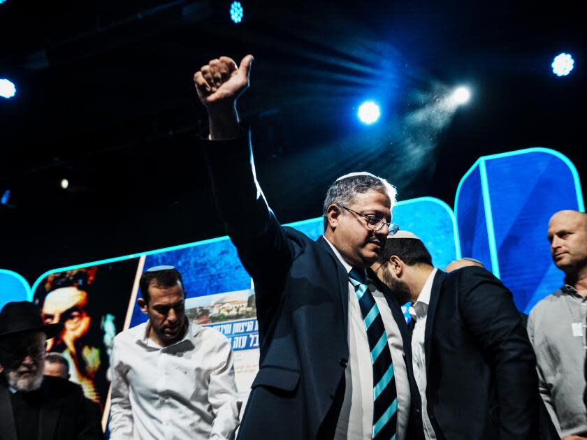 Itamar Ben-Gvir gives a thumbs-up and hugs a man as others stand by on a stage decorated with blue panels