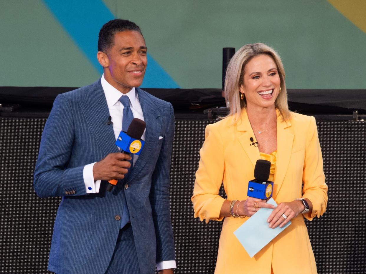 T.J. Holmes, in a blue suit, smiles while standing next to fellow "GMA" host Amy Robach, in a yellow blazer and skirt, on stage.