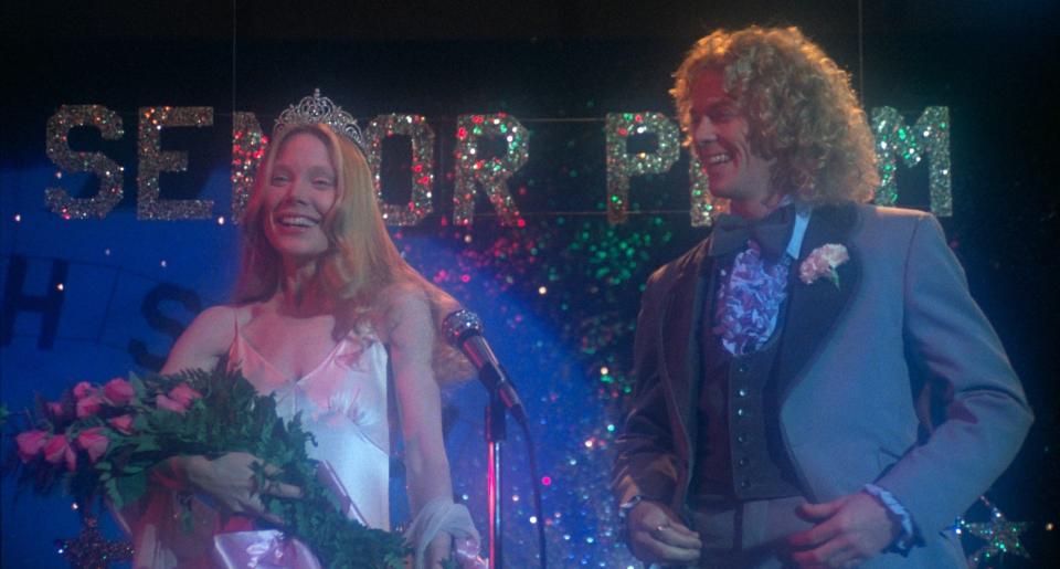 Sissy Spacek, wearing a gown and holding roses, and William Katt, in a tuxedo, are on a stage labeled "Senior Prom." Sissy wears a tiara and smiles at the mic