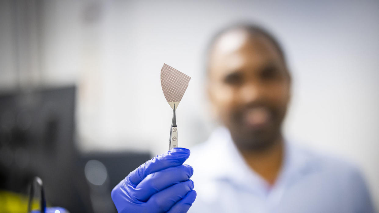  A photo of a material science researcher holding a piece of AlScN-based flash memory in tweezers, with the person out of focus in the background. 