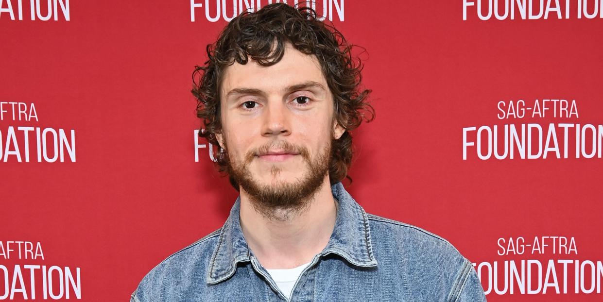 evan peters with a beard wearing a denim shirt and smiling in front of a red background