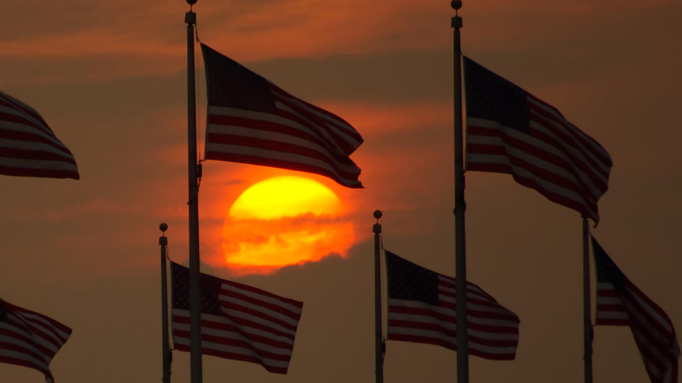  An orange sun sets behind a wall of clouds as several american flags wave in the foreground. 