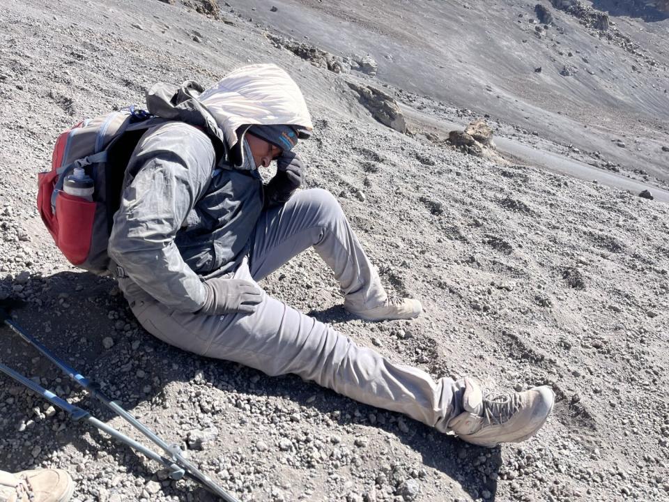 A woman in a light gray jacket and pants and carrying a backpack holds a hand to her forehead as she sits on gray soil