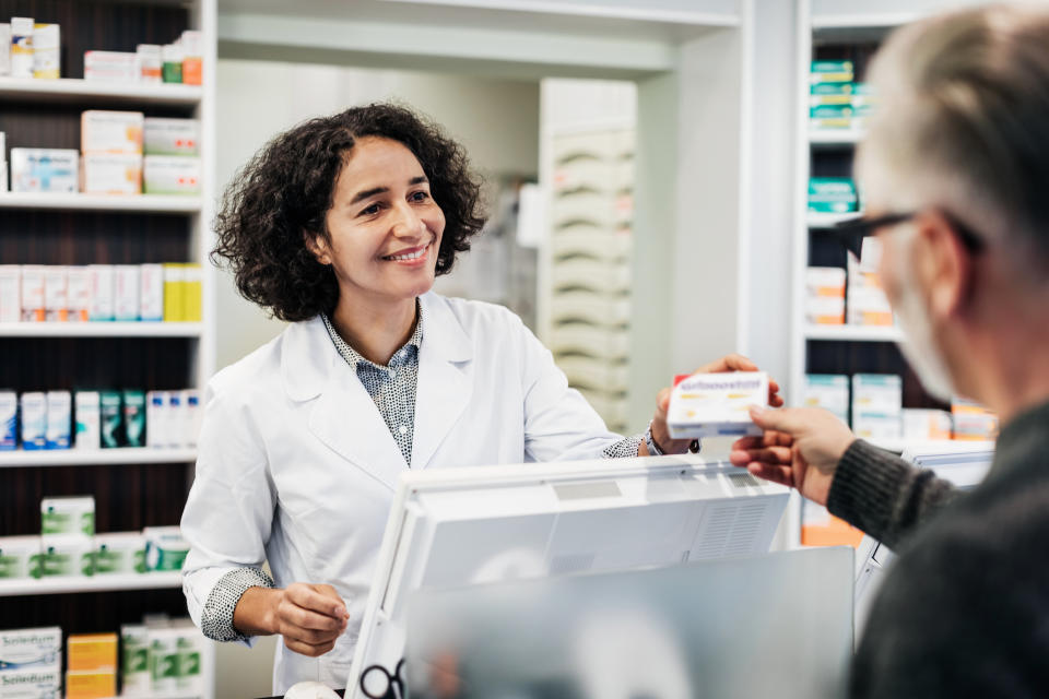 Pharmacist smiling and handing a medication to a customer across the counter in a pharmacy setting. Shelves with various medicines are visible in the background