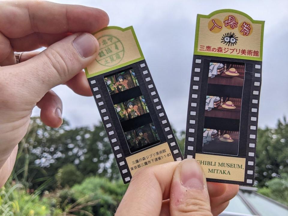 Close-up of film tickets that show negatives from Studio Ghibli films held up against the cloudy sky. The one on the right shows a hat on a wooden bench, next to the corner of a woman's dress. The one on the left shows a man dressed in a red cape and green shirt.