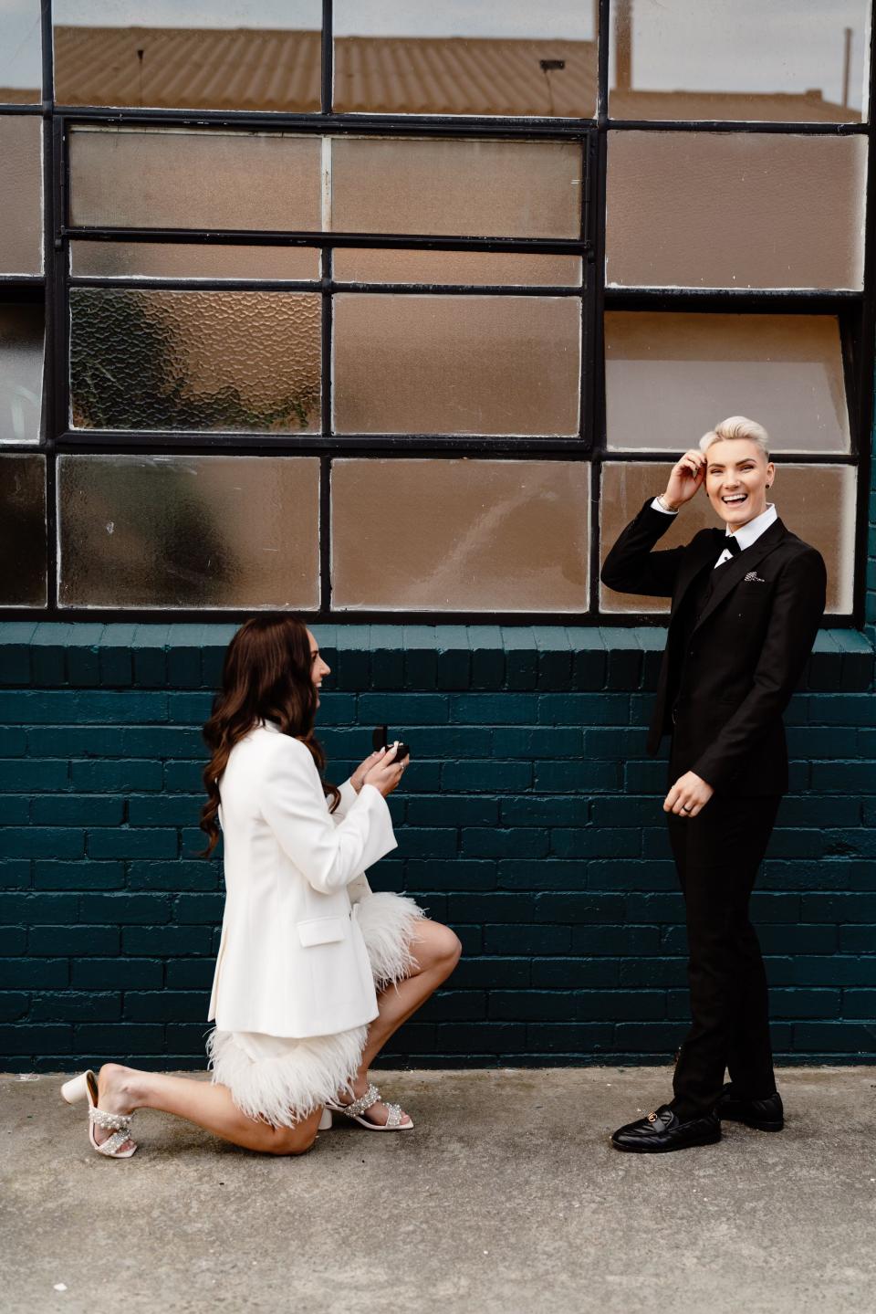 Morgan Owen proposing to her partner Axe. She's wearing a white dress and down on one knee.