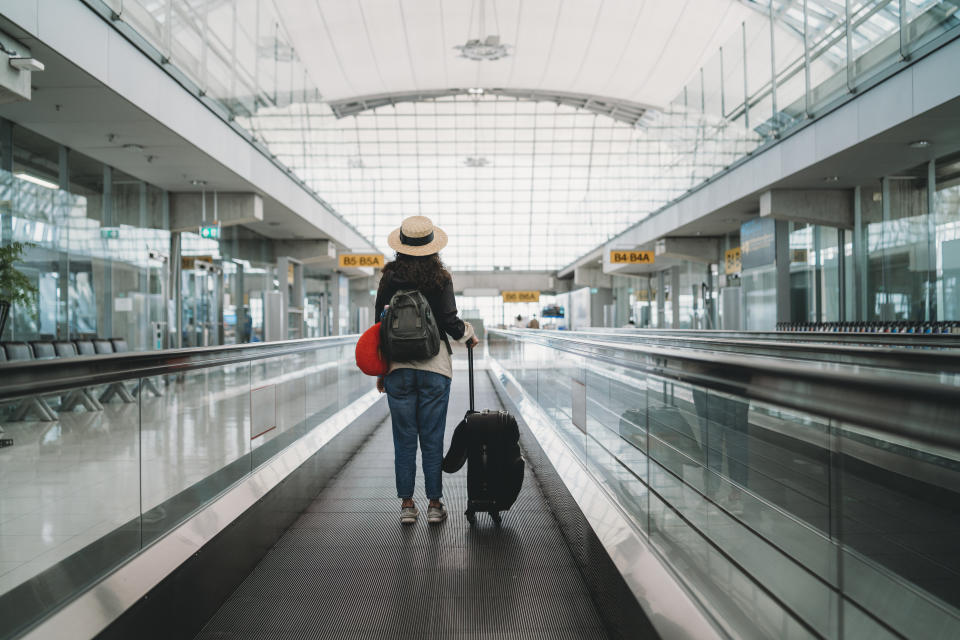 A tourist wearing a hat has her back to the camera as she rides a travelator in an airport. 