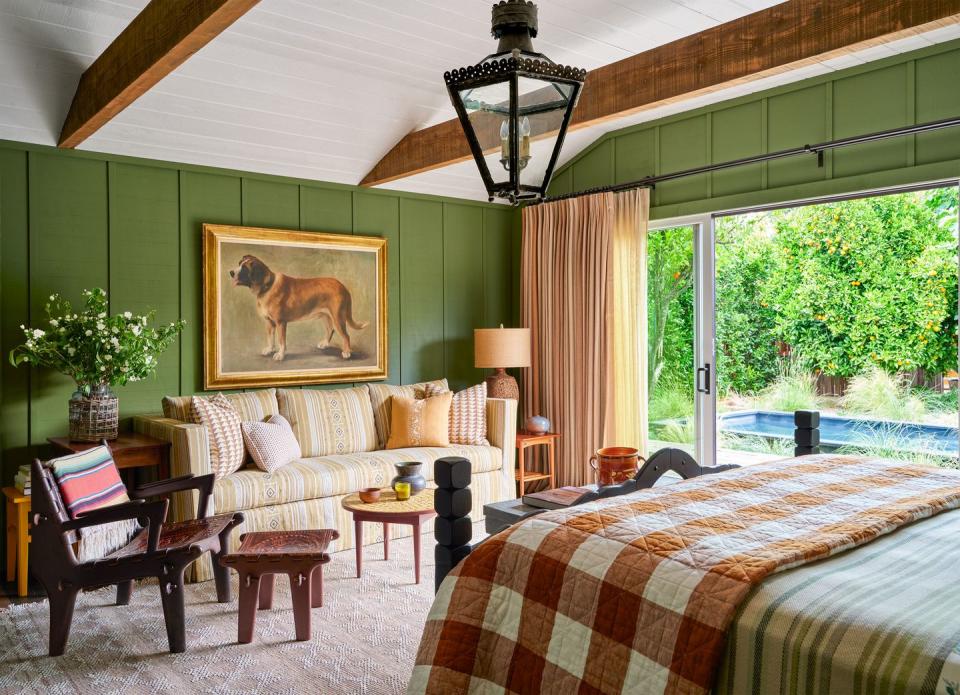 green walls, a light patterned rug, striped fabric sofa with framed portrait of a dog above, wood armchair with striped blanket, green striped bed cover and brown and white plaid quilt, curtains on sliding glass doors to pool