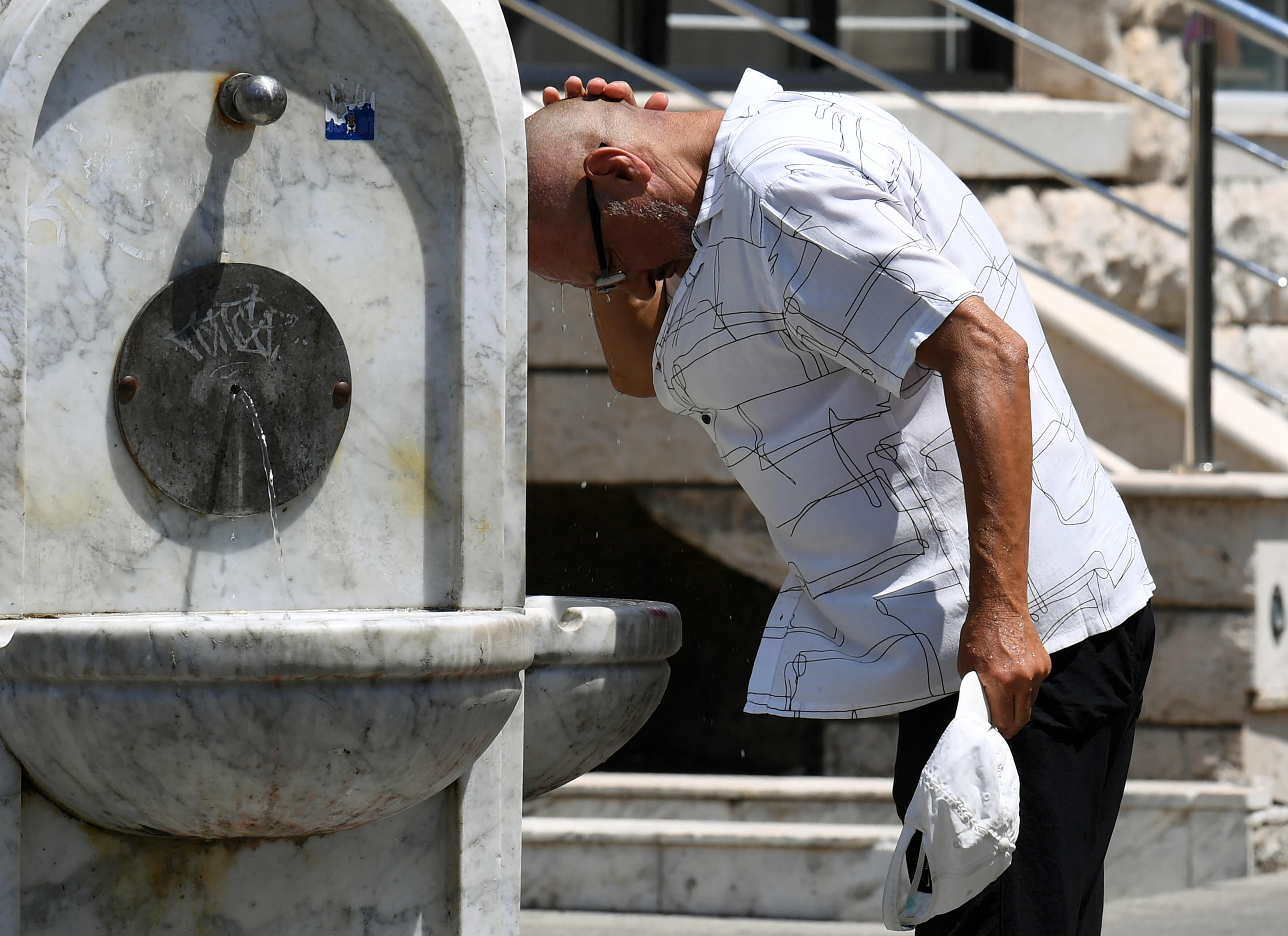 A pedestrian cools his head by running it under a public water tap on a street.