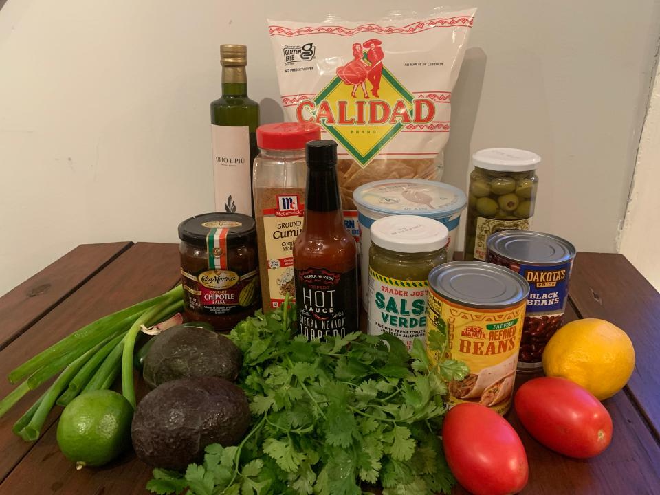 A cutting board of ingredients including avocado, lime, lemon, tomatoes, salsas, beans, tortilla chips, and scallions