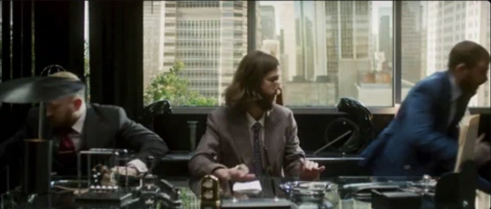 Three people in business attire are seated at a glass table in an office with a cityscape view. The person on the right is moving quickly
