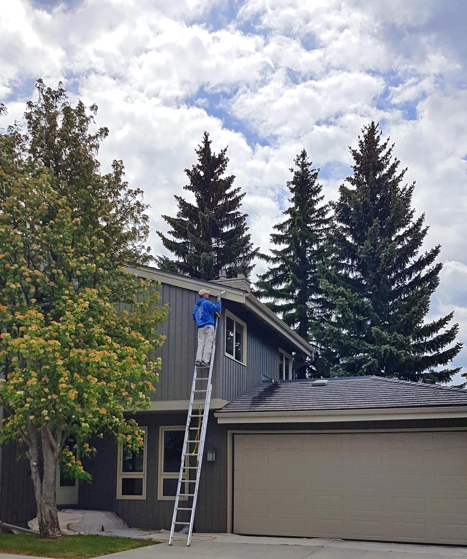 Person standing on a tall ladder working on the roof of a two-story house surrounded by trees