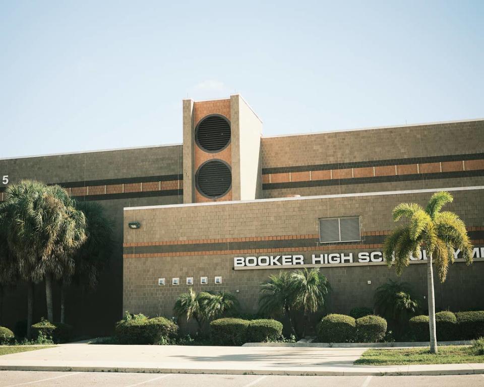 A brown, brick school with lighter brown accents. A sign on the school reads "BOOKER HIGH SCHOOL" in a wide sans-serif font. There are palm trees around the school. The building is very dimensional.