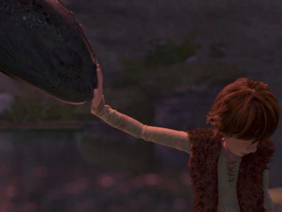 a teenage boy (right) in a green shirt and brown vest reaches his hand out to a pitch black dragon on the left. the dragon is butting its nose against the boy's hand and they're both closing their eyes.