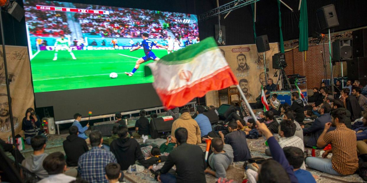 An Iranian flag flies as fans sit in front of a screen and watch the US-Iran World Cup match.
